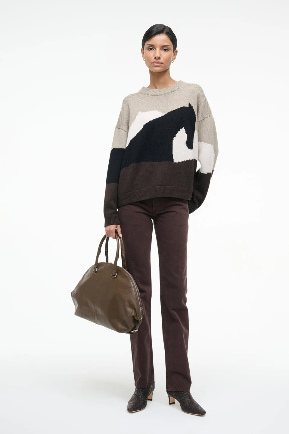 Woman wearing a color-blocked sweater and brown pants, holding a green bag on a white background