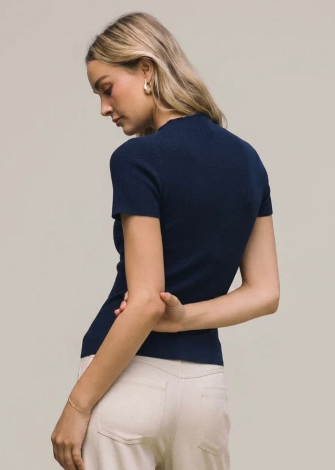 A woman with blonde hair is shown from the back wearing a navy blue short-sleeve knit top and cream-colored pants, set against a plain light gray studio background.