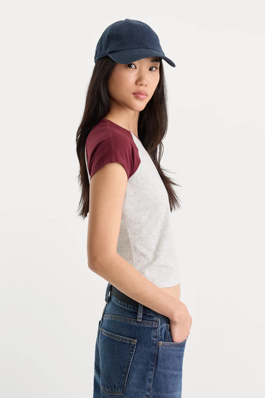 A model wearing a short-sleeve raglan t-shirt with a light gray body and dark burgundy sleeves, paired with a navy blue baseball cap and blue jeans, shown in profile against a plain white studio background.