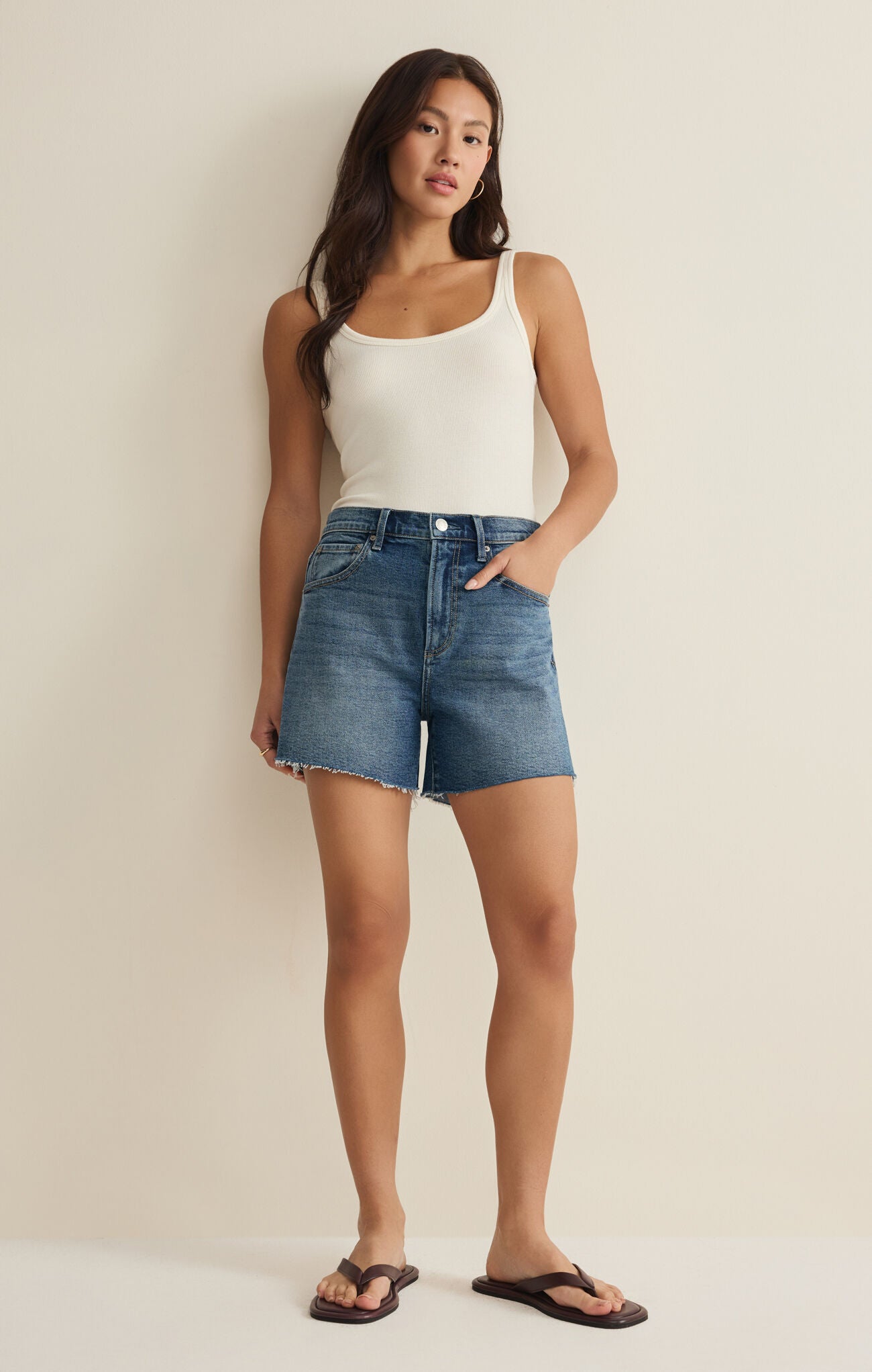 Woman wearing a white tank top and blue denim shorts against a beige background