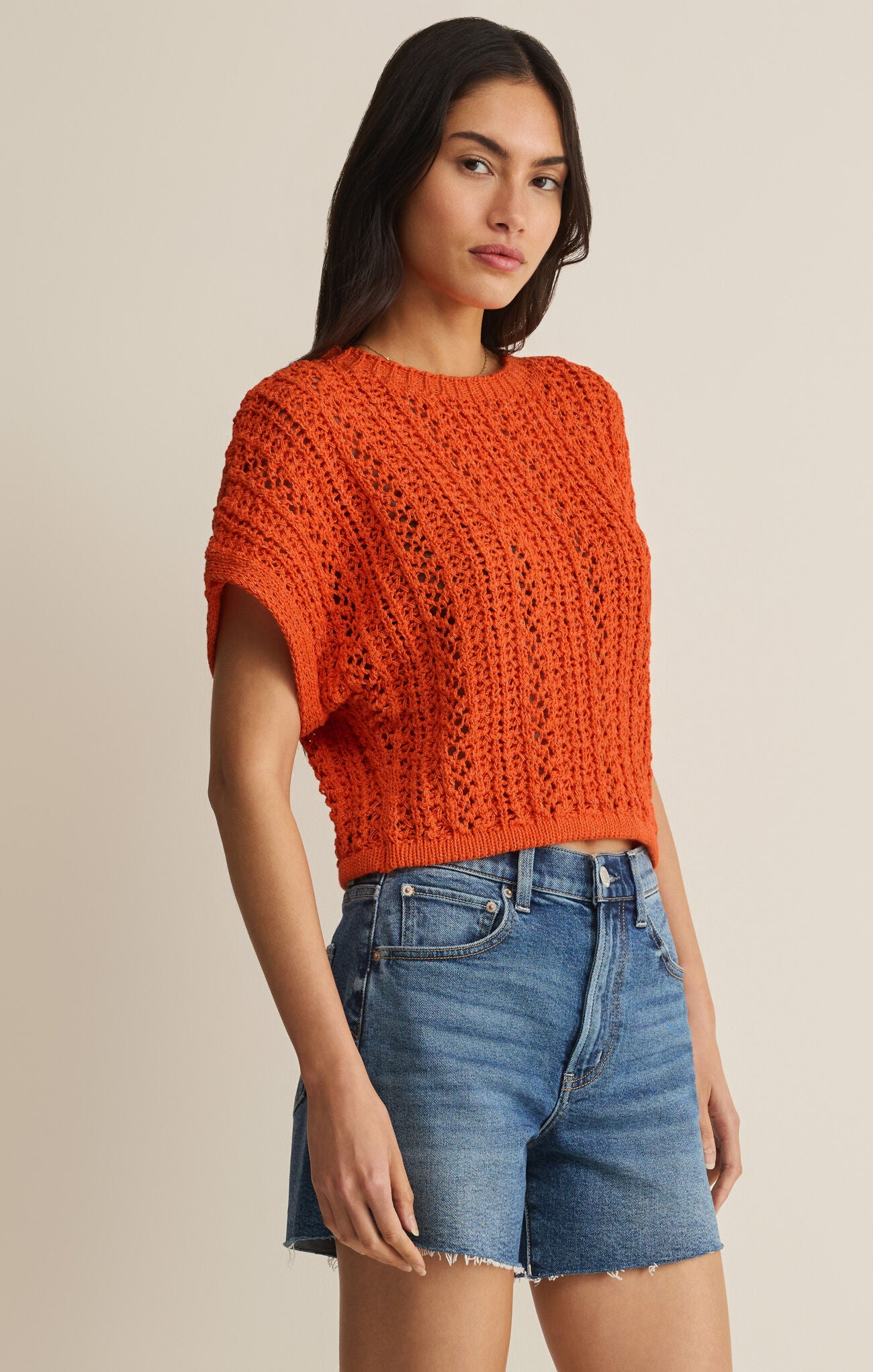 Woman wearing an orange knitted top and denim shorts on a beige background