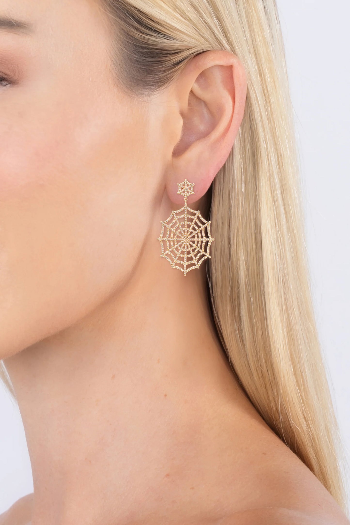 A close-up of a woman's ear wearing a delicate gold spiderweb dangle earring against a clean white background.