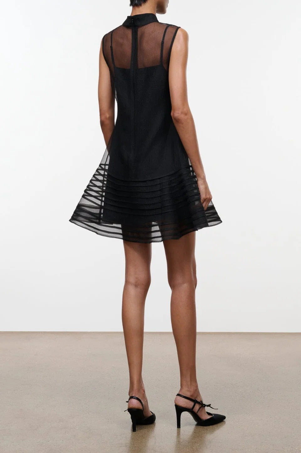 Model on a neutral background wearing the STAUD sterling mini dress in black which features a sheer outer layer with a high neck and a mini black slip underneath. The back view showcases the button closure on the high neck.