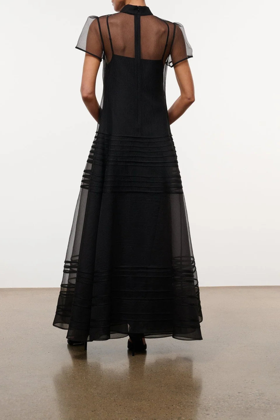 Model wearing the STAUD Stratton maxi dress that features a sheer outer layer with a black maxi slip underneath. The outer layer features a mock neckline and capped sleeves. The back view shows the button closure on the mock neckline.