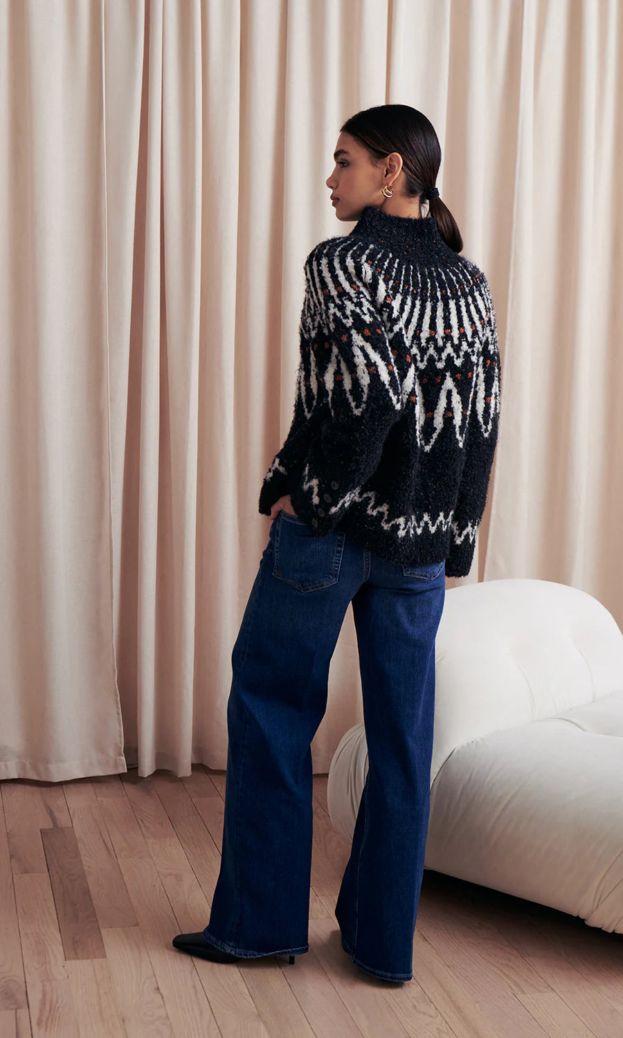 Person wearing a black and white patterned sweater and blue jeans standing in a room with beige curtains.