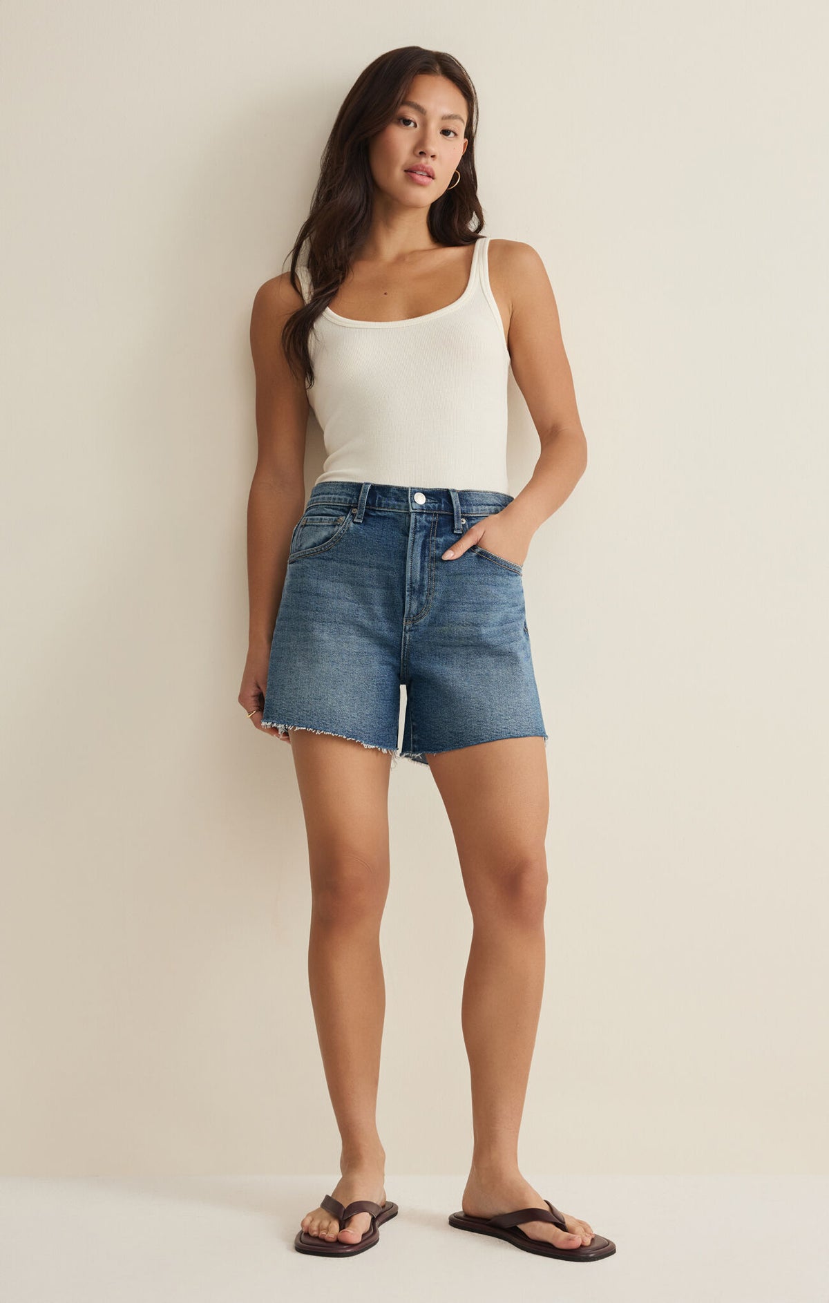 Woman wearing a white tank top and blue denim shorts against a beige background
