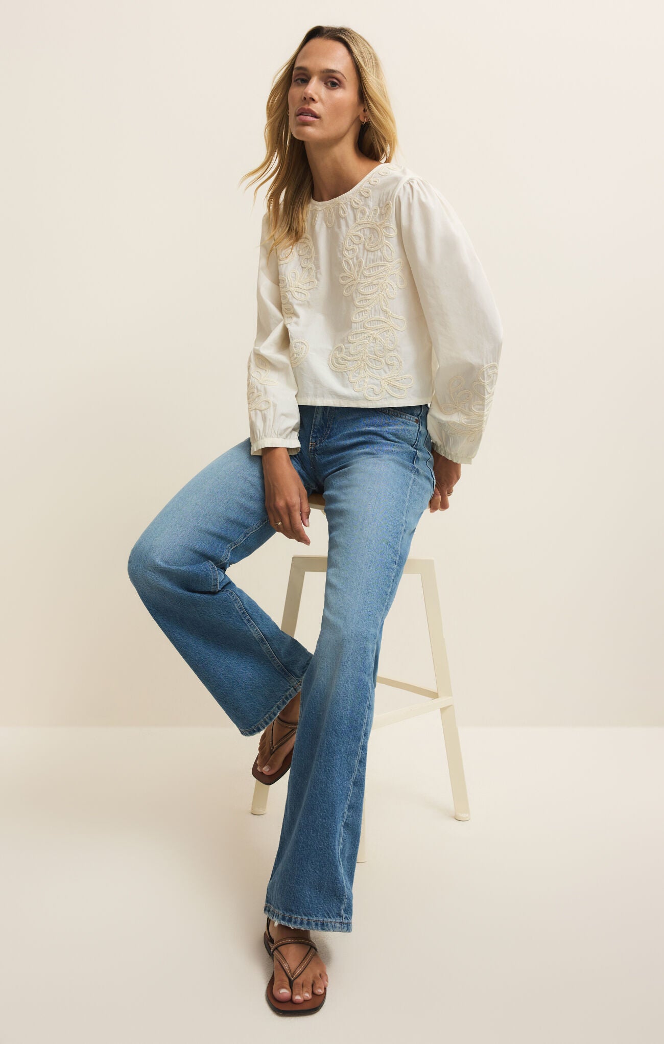 Woman wearing a white sweater and blue jeans sitting on a white stool against a plain background