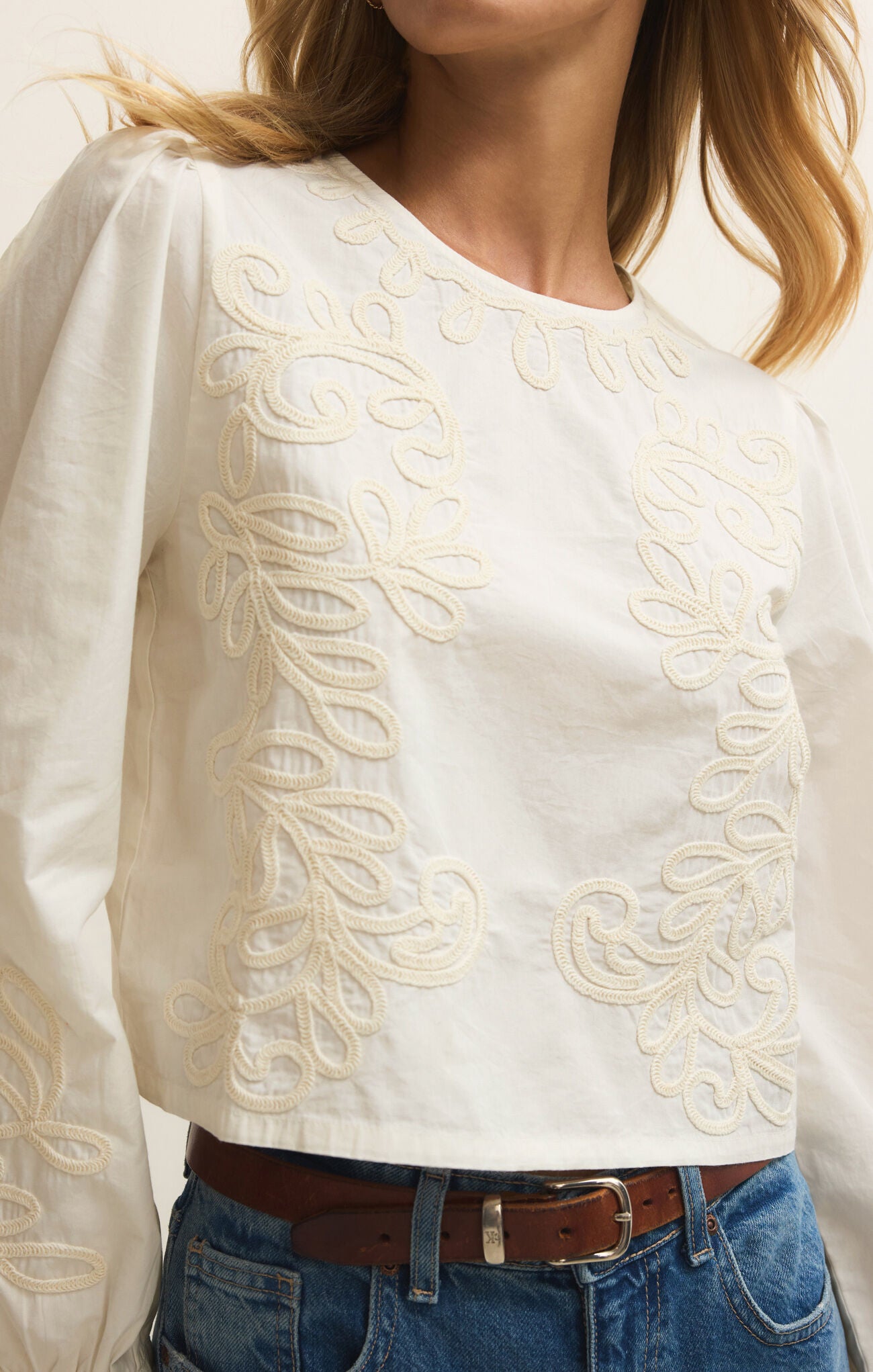 White blouse with intricate embroidery worn by a person, paired with blue jeans and a brown belt.