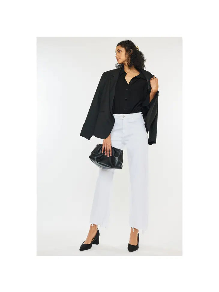 Woman wearing white high rise denim jeans with a frayed bottom hem wearing a black polo, blazer, high heels, and holding a black bag.