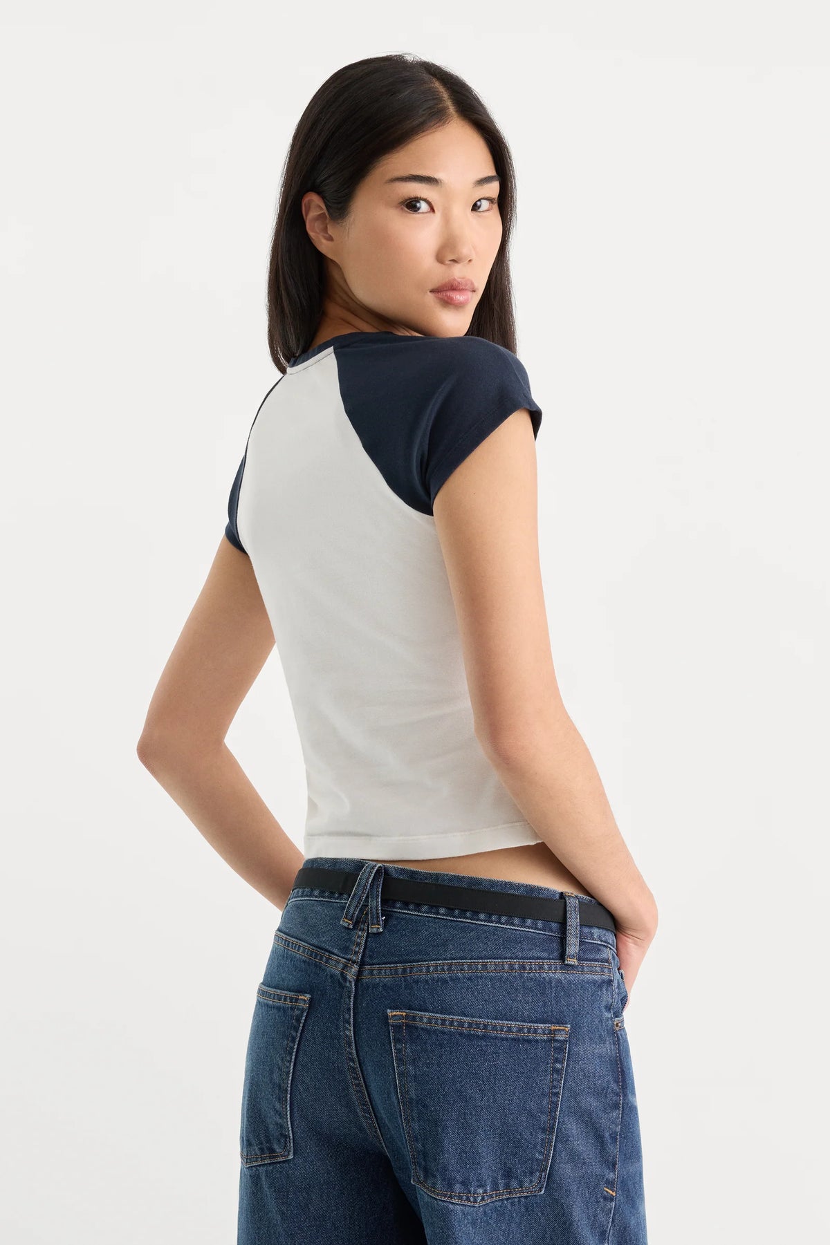 A woman wearing a short-sleeve raglan t-shirt with a white body and navy blue sleeves, paired with blue jeans, photographed from the back against a plain white studio background.
