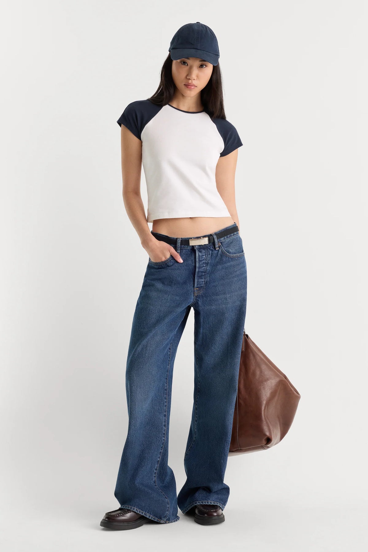 A model stands against a light gray studio background wearing a white and navy raglan crop top, blue wide-leg jeans, a navy cap, and holding a brown leather bag.