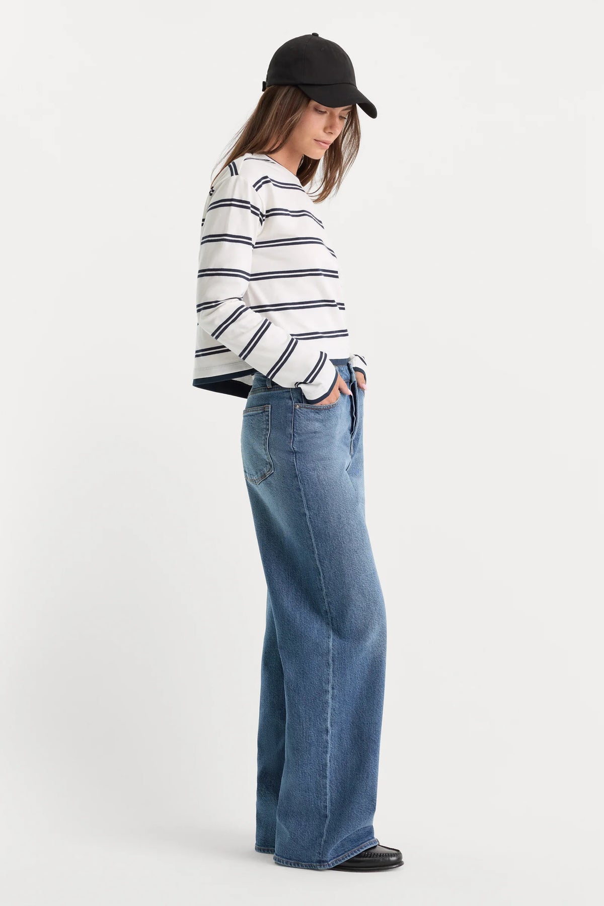A model wearing a white long-sleeve top with thin navy horizontal stripes, high-waisted wide-leg blue jeans, and a black baseball cap, standing against a plain white studio background.