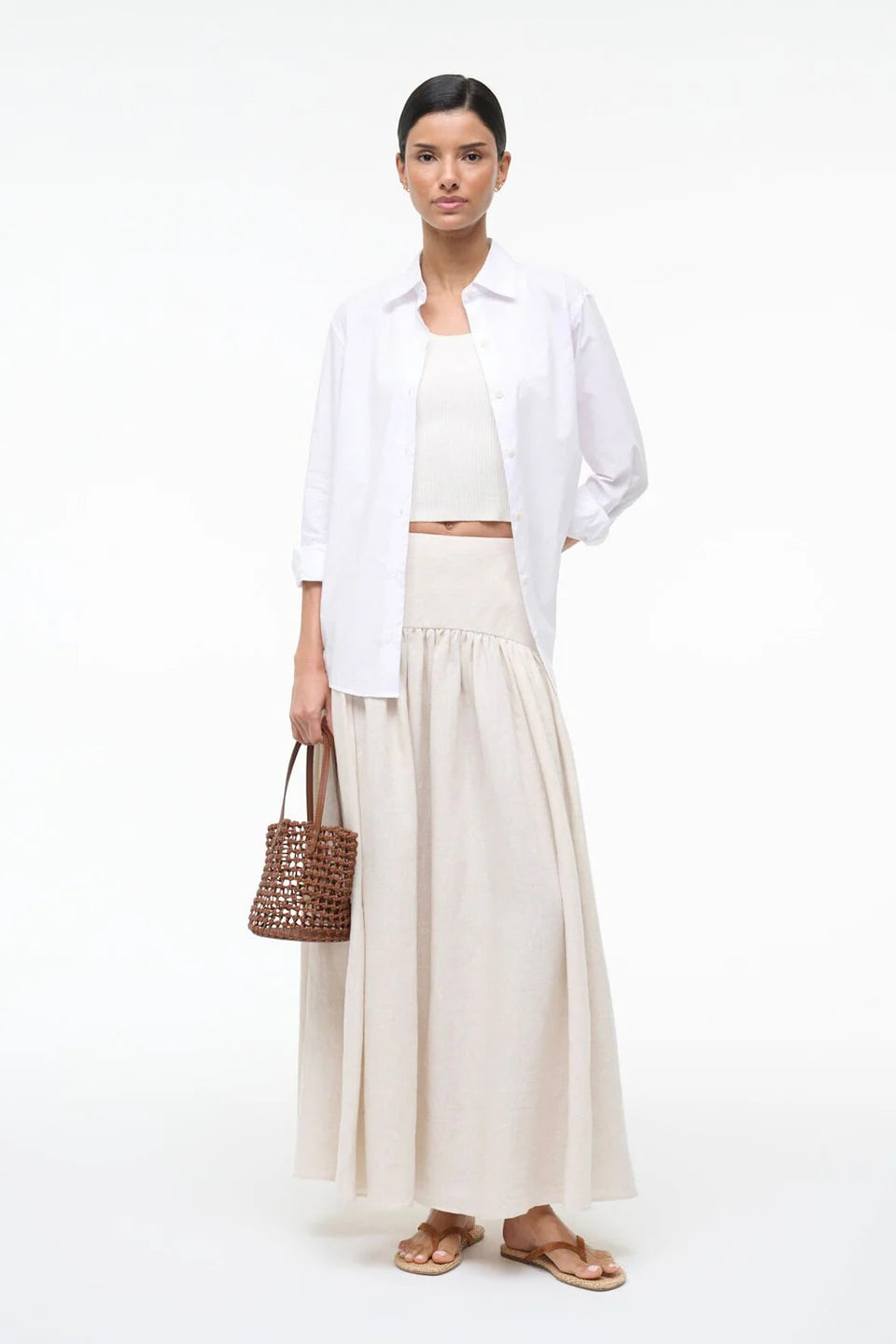 Woman wearing a white shirt, beige skirt, and holding a woven bag on a white background