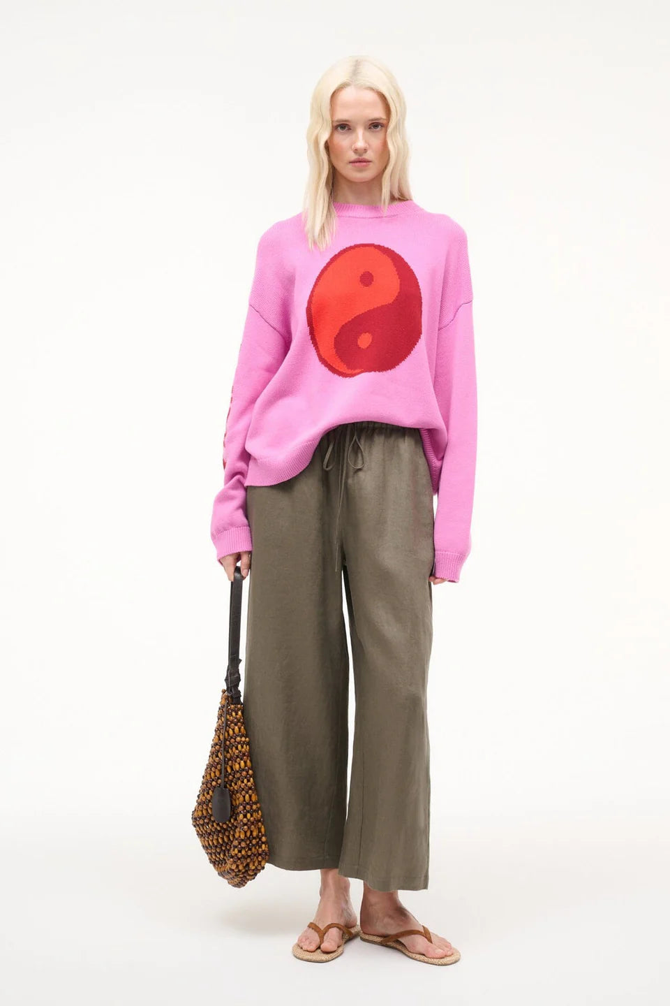 Person wearing a pink sweater with a red yin yang symbol, holding a patterned bag on a white background