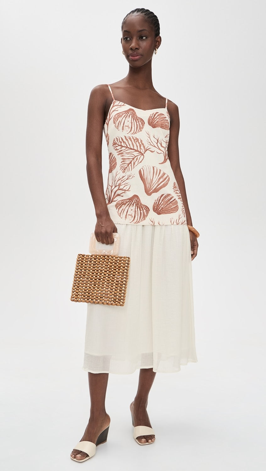 Woman wearing a sleeveless top with shell pattern and white skirt holding a woven clutch.