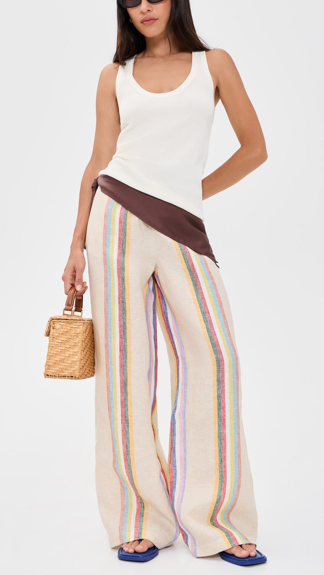 Woman wearing a white sleeveless top and rainbow-striped pants, holding a straw bag on a white background
