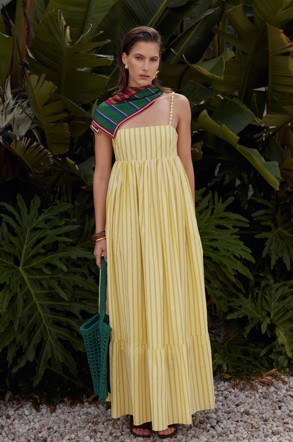 Woman in a yellow dress with a colorful top standing in front of green foliage.
