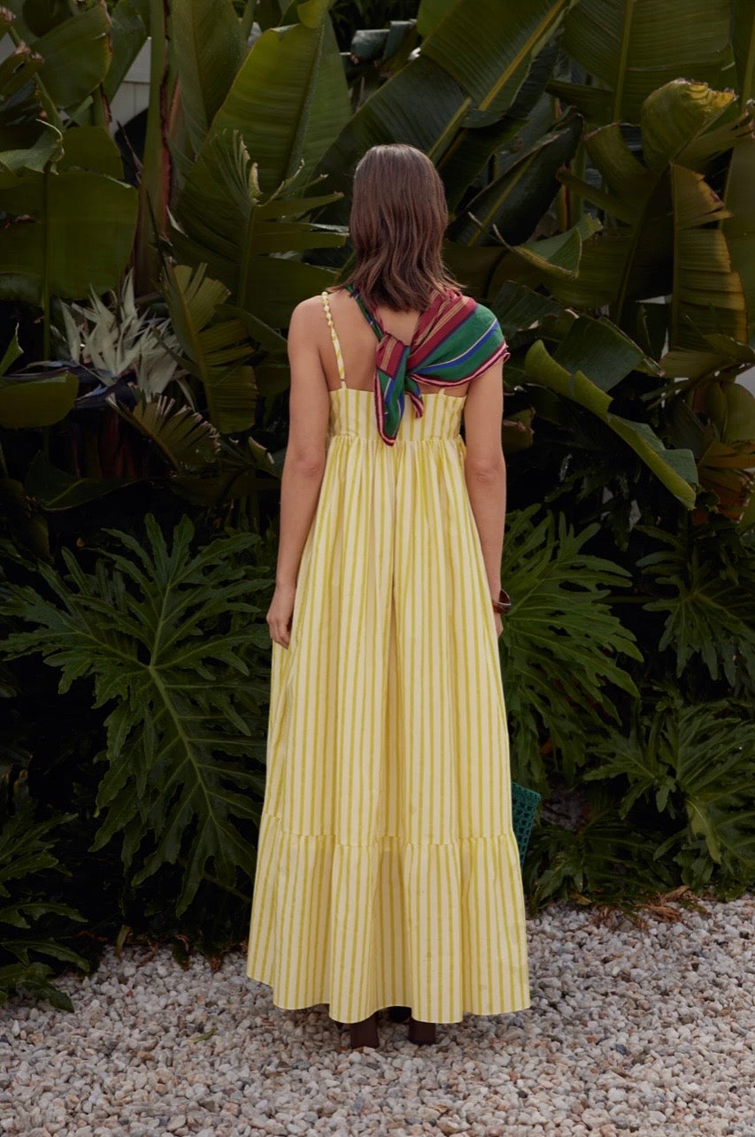 Woman in a yellow dress with a colorful scarf standing in front of green foliage.