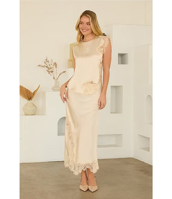A woman modeling a cream satin slip dress with lace trim, standing in a minimalist white room with geometric wall shelves.
