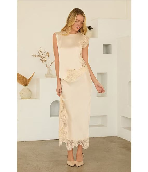 A woman models a cream satin midi dress with lace trim details, standing in a minimalist white room with geometric shelves.