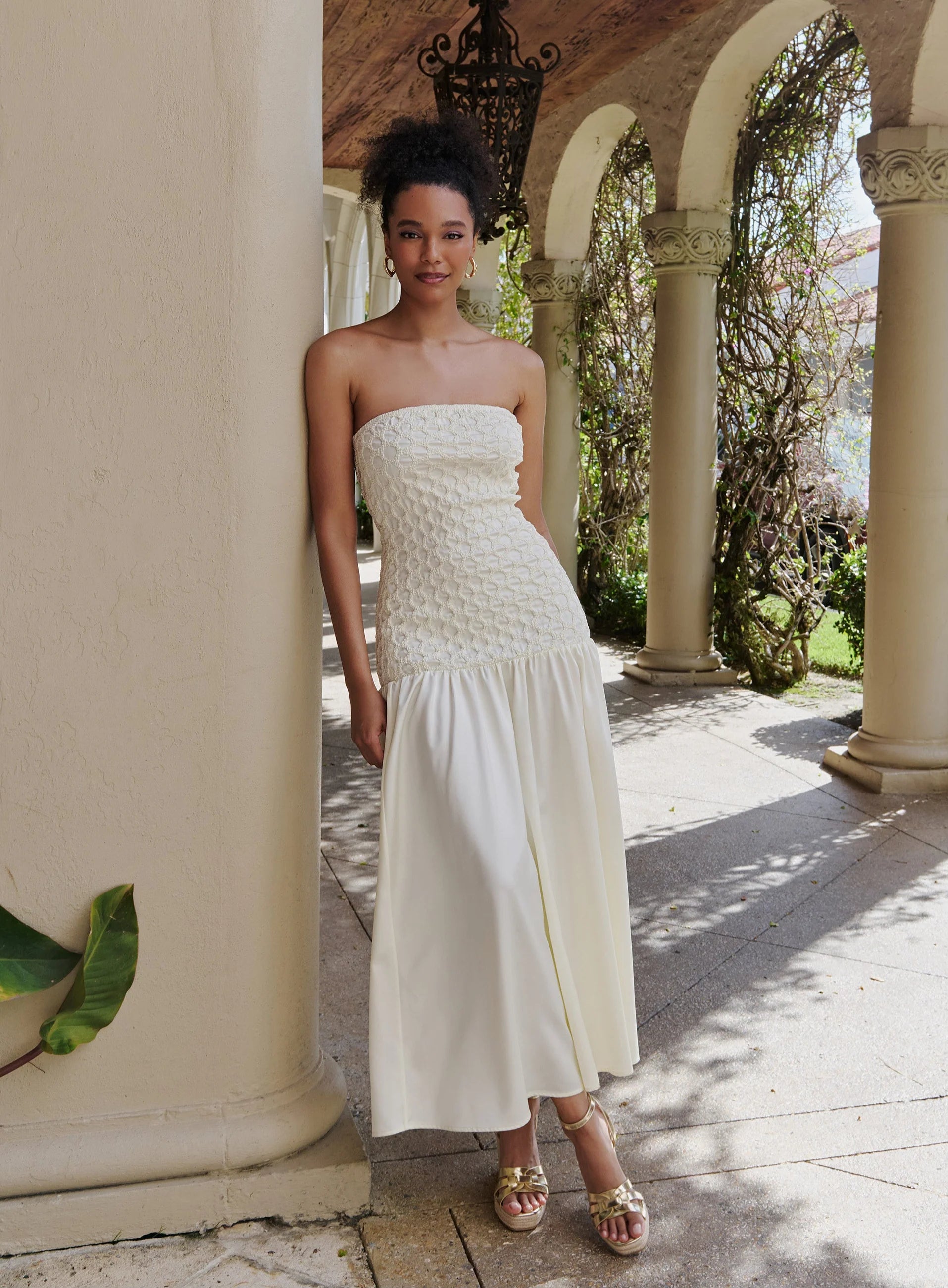 A woman models a strapless white maxi dress with a textured crochet bodice and a flowing skirt, standing in a sunny outdoor colonnade with stone columns and arches.