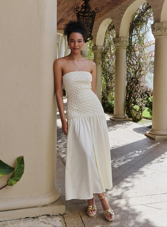 A woman models a strapless white maxi dress with a textured crochet bodice and a flowing skirt, standing in a sunny outdoor colonnade with stone columns and arches.