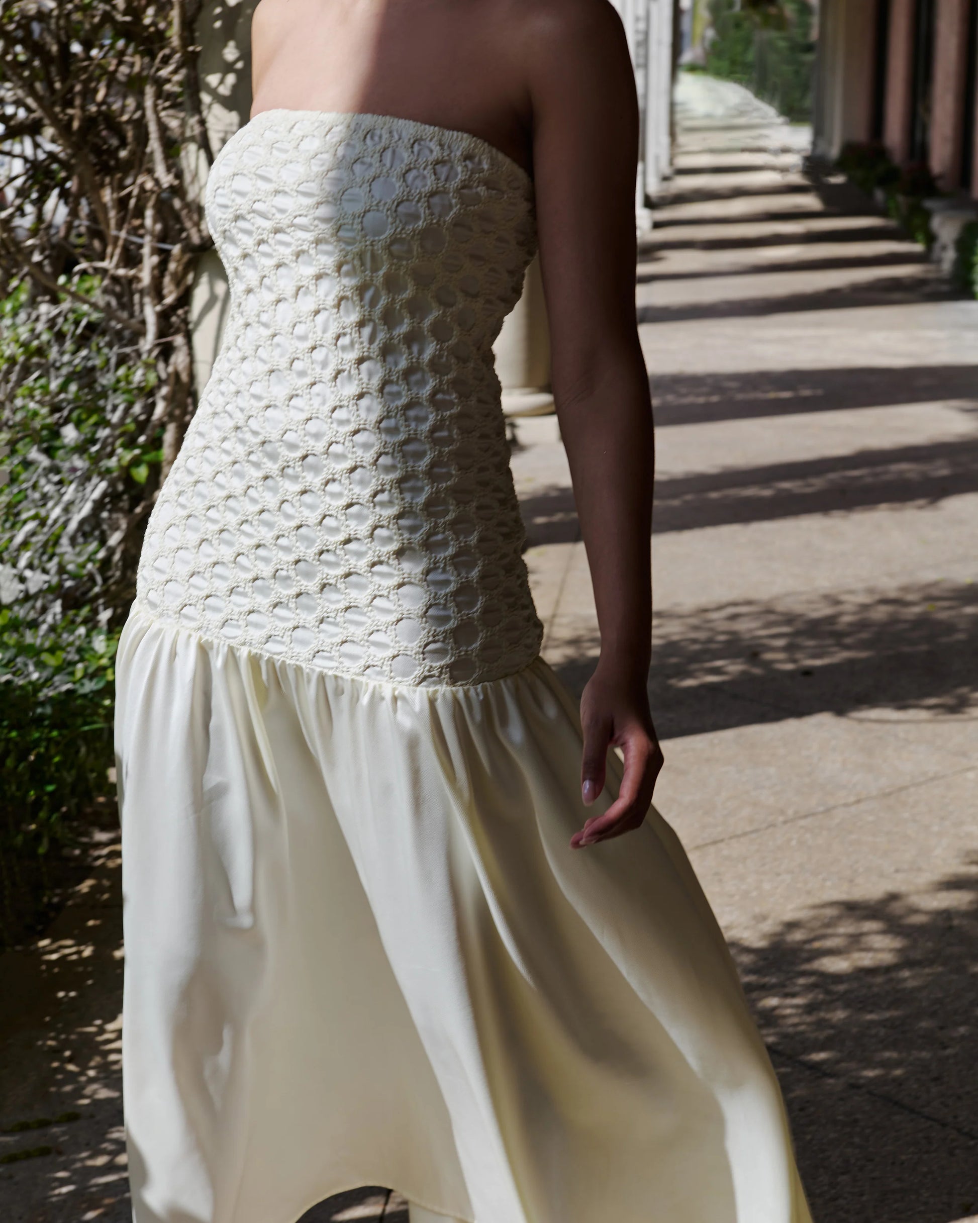 A woman wearing a strapless cream dress with a heavily textured, patterned bodice and a flowing satin-like skirt, photographed outdoors on a sunlit walkway.