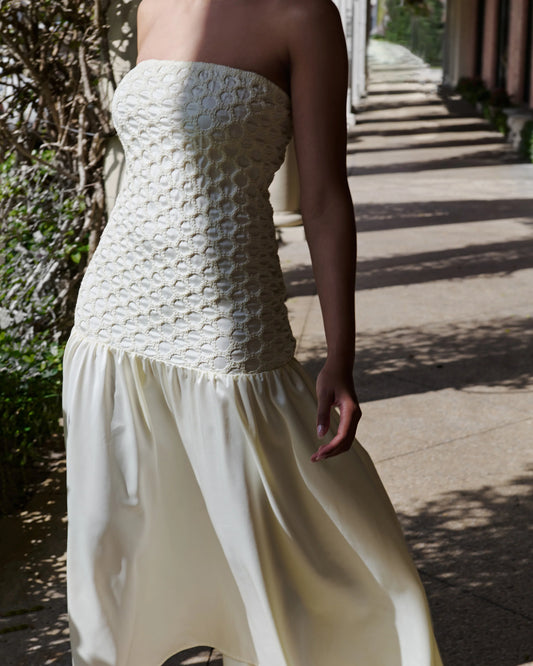 A woman wearing a strapless cream dress with a heavily textured, patterned bodice and a flowing satin-like skirt, photographed outdoors on a sunlit walkway.