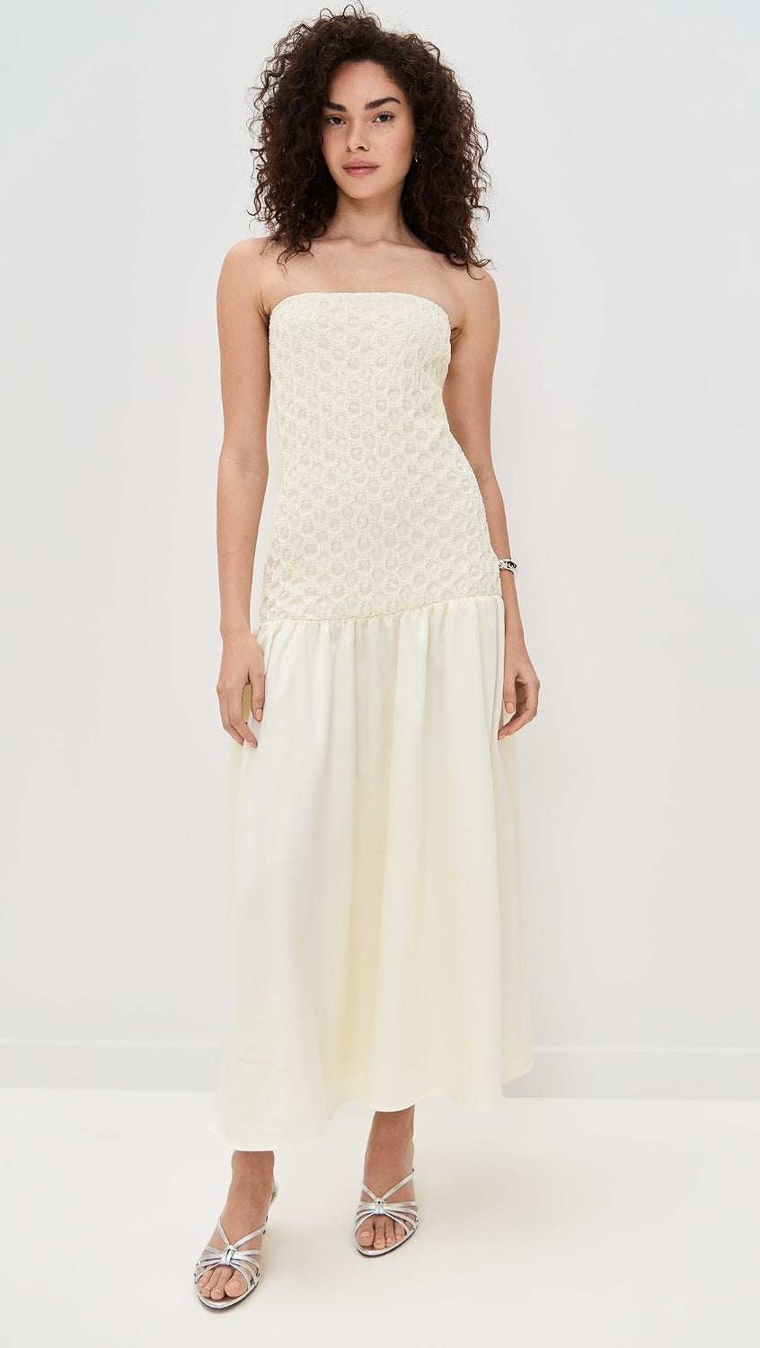 A woman modeling a strapless cream maxi dress with a textured bodice and a flowing skirt, paired with silver strappy heels, against a plain white studio background.