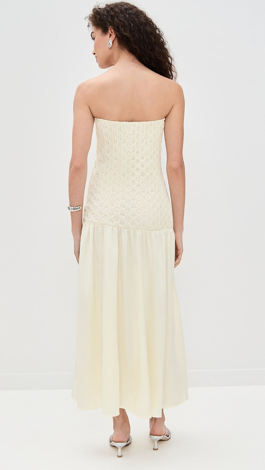A woman shown from the back wearing a strapless cream maxi dress with a textured bodice and flowing skirt, paired with silver high-heeled sandals against a plain white studio background.