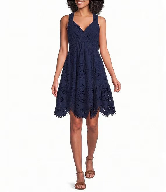 A woman modeling a sleeveless navy blue eyelet lace mini dress with a gathered bodice and flared skirt, paired with brown strappy sandals against a plain white background.