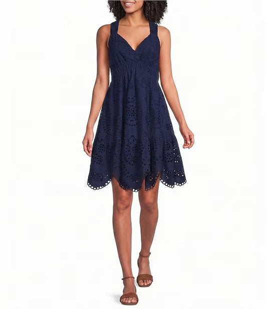 A woman modeling a sleeveless navy blue eyelet lace mini dress with a gathered bodice and flared skirt, paired with brown strappy sandals against a plain white background.