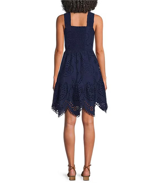 A woman shown from the back wearing a sleeveless navy blue eyelet lace dress with a square neckline and a flared skirt, paired with brown heeled sandals against a plain white background.