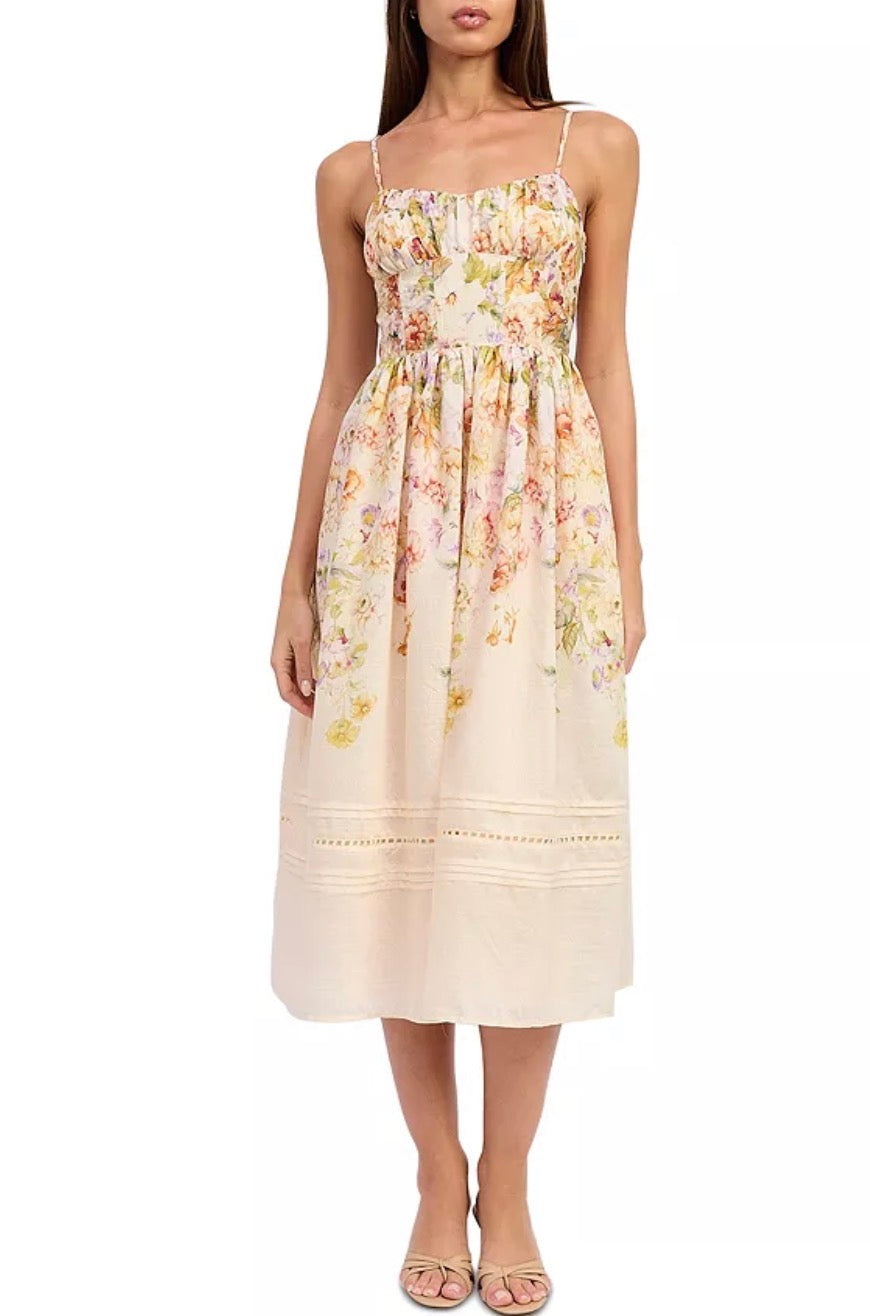 A woman modeling a sleeveless midi dress with thin straps, a smocked bodice, and a soft floral print in pastel tones on a cream base, photographed against a plain white studio background.