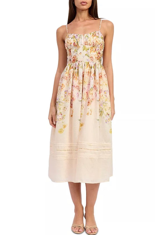 A woman modeling a sleeveless midi dress with thin straps, a smocked bodice, and a soft floral print in pastel tones on a cream base, photographed against a plain white studio background.