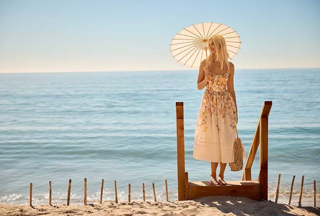 A woman stands on a wooden platform by the beach holding a cream-colored paper umbrella. She is wearing a sleeveless, floral-printed midi dress and holding a small woven bag.