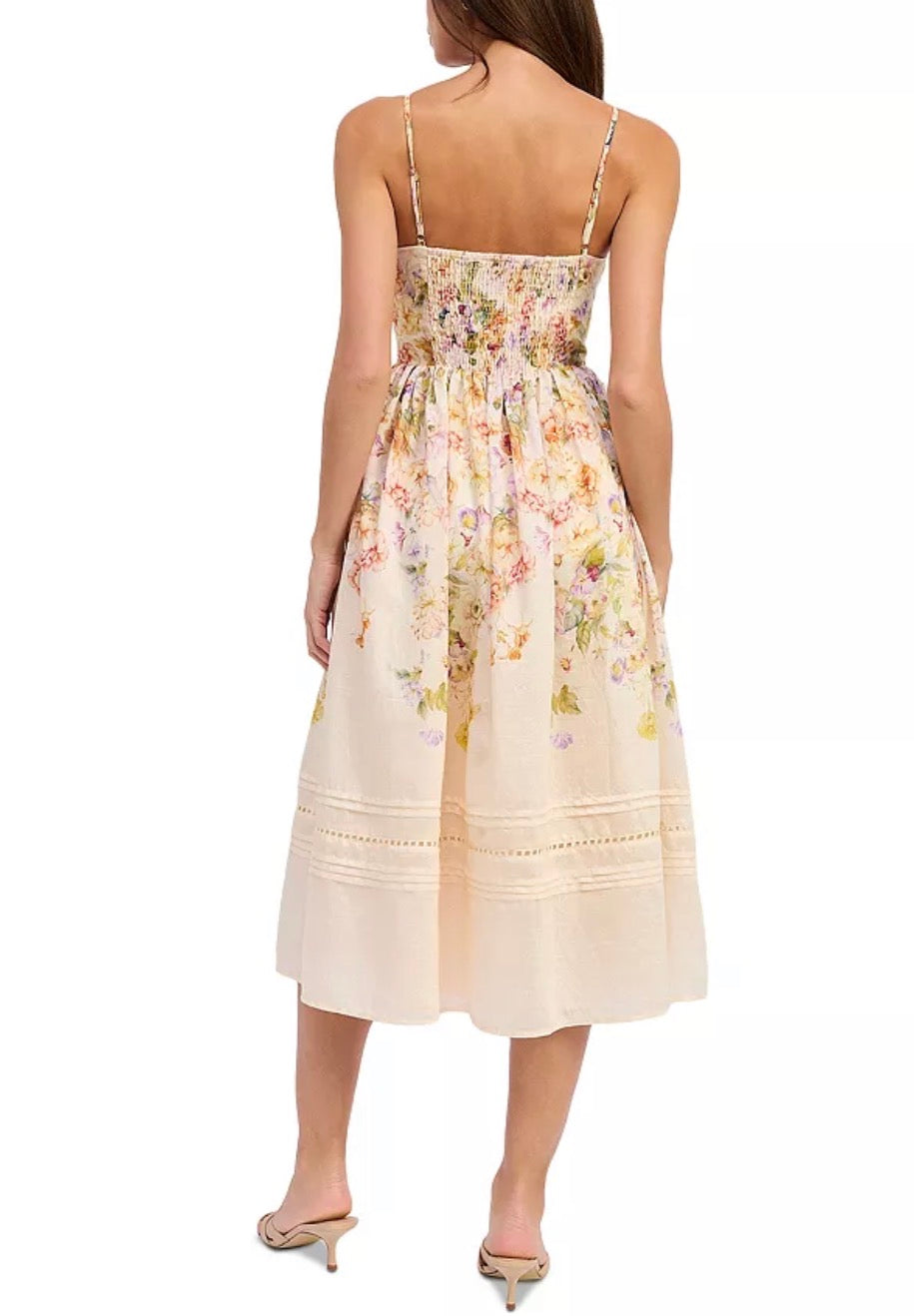 A woman modeling a sleeveless midi dress from the back, featuring a cream base with a soft pastel floral print and delicate tie-shoulder straps, set against a plain white background.