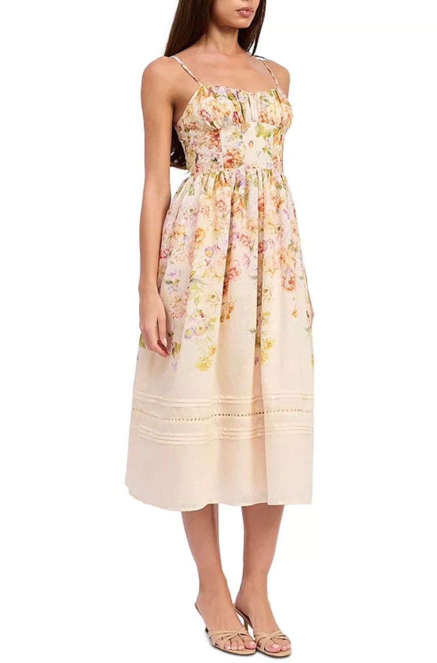 A woman modeling a sleeveless midi dress with thin straps, a gathered bodice, and a soft floral print in pastel tones on a light cream base. The dress has embroidered detailing near the hem and is shown against a plain white background.