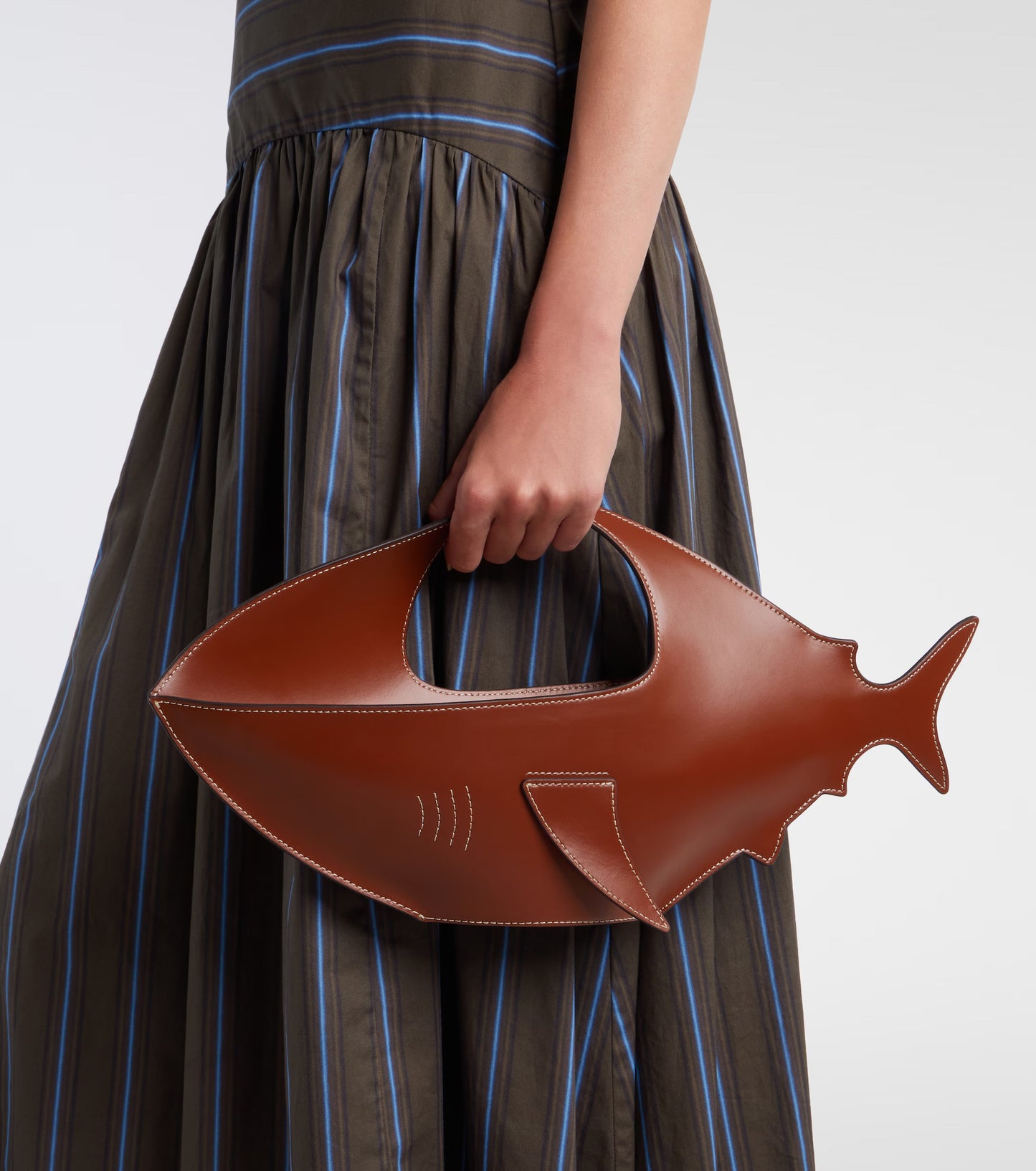 A person in a brown and blue striped dress holds a brown leather fish-shaped handbag with a handle cutout, shown against a plain light gray background.
