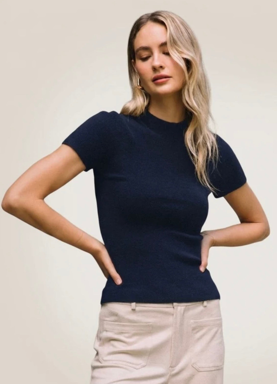 A woman modeling a short-sleeve, crew-neck knit top in dark navy blue, paired with light beige pants, against a plain light beige studio background.