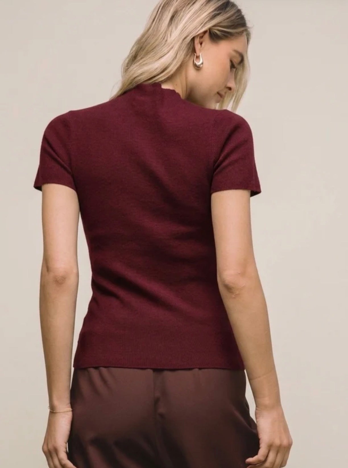 A woman shown from the back wearing a fitted short-sleeve knit top in deep burgundy, paired with dark brown bottoms, against a plain light beige studio background.