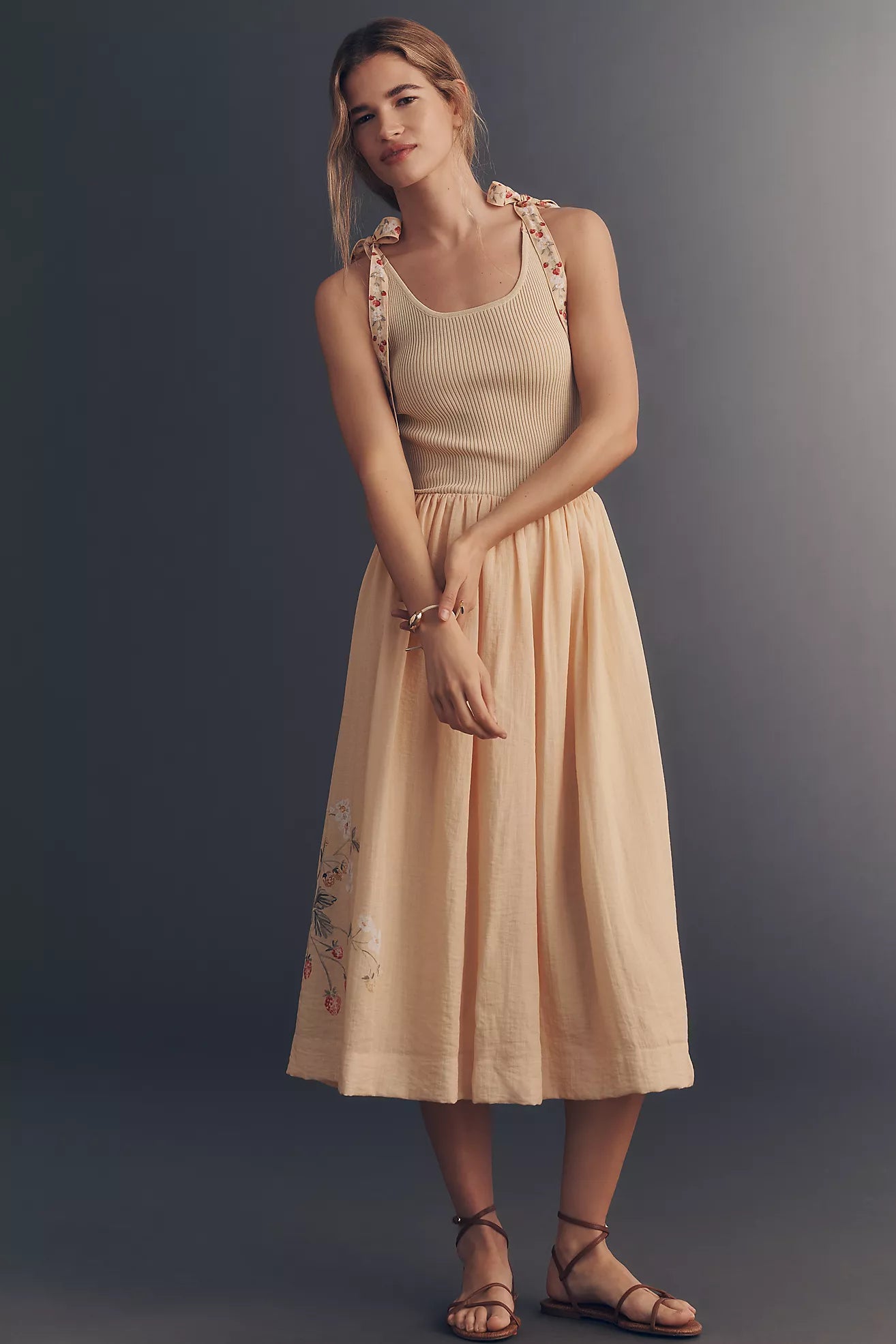 A woman modeling a sleeveless midi dress in a light peach-beige color, featuring a ribbed knit bodice, floral embroidered shoulder straps, and matching floral embroidery on the skirt, paired with brown strappy sandals against a dark gray studio background.