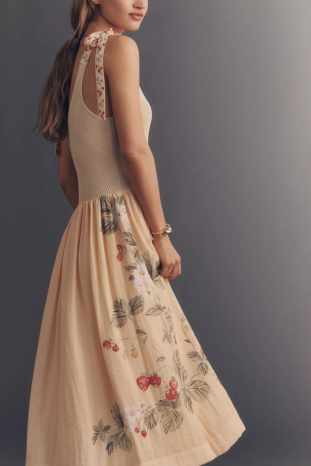 A woman wearing a sleeveless beige maxi dress with a ribbed knit bodice and a flared skirt featuring a delicate floral and berry print, shown against a dark gray studio background.