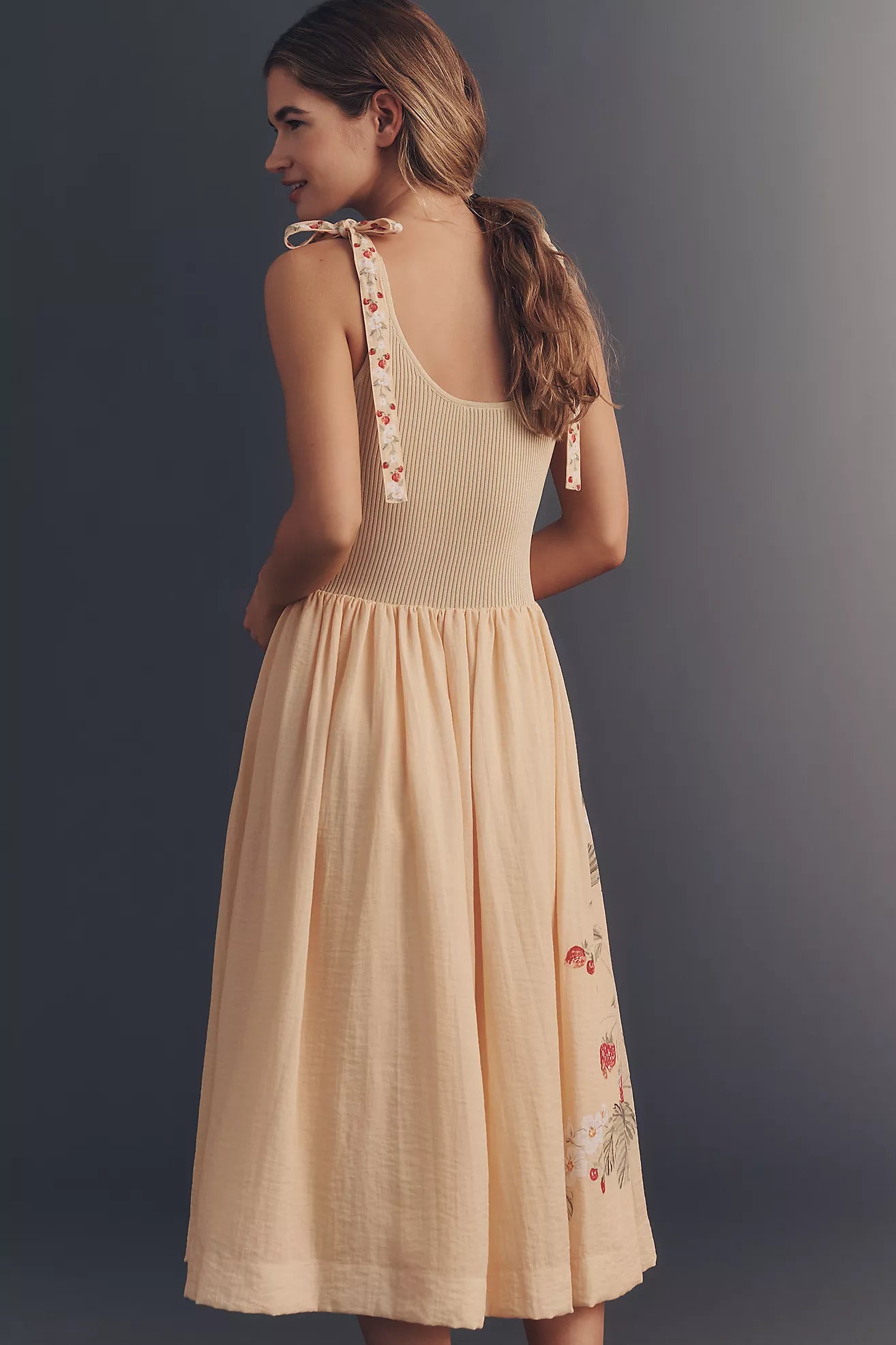 A woman shown from the back wearing a sleeveless beige dress with tie straps and floral embroidery near the hem, photographed against a dark gray studio background.