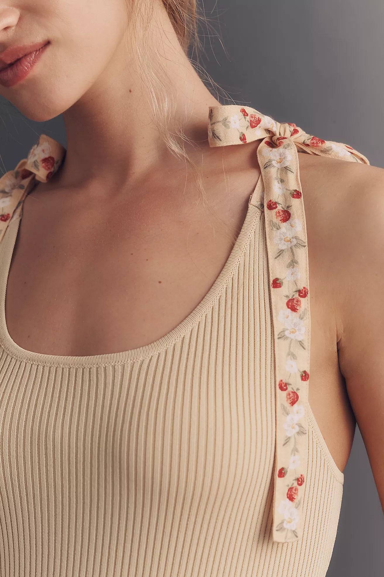 Close-up of a woman wearing a beige ribbed tank top with floral ribbon shoulder ties in a cherry print, against a dark gray background.
