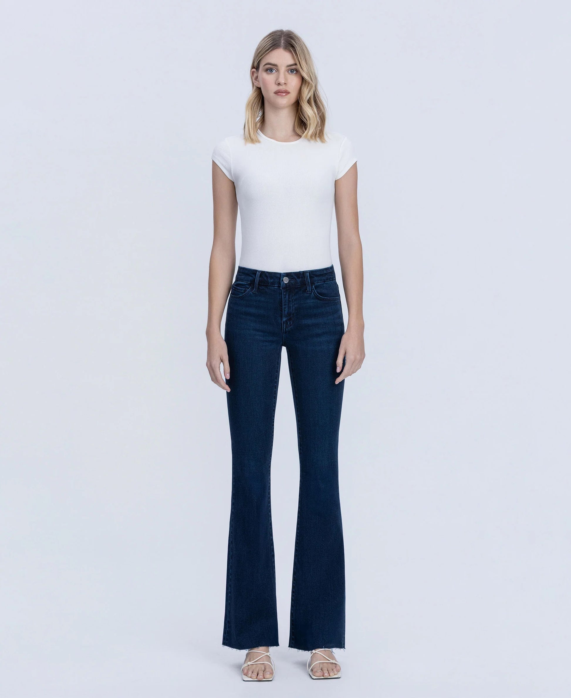 A woman modeling dark blue flare jeans with a white short-sleeve top, standing against a light gray studio background.