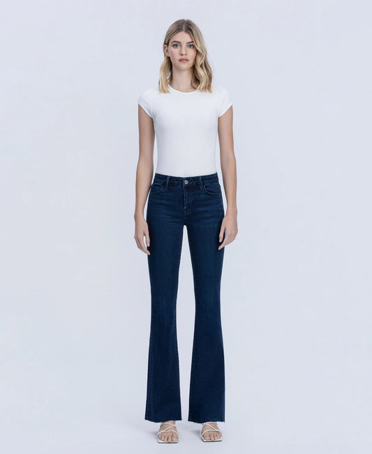 A woman modeling dark blue flare jeans with a white short-sleeve top, standing against a light gray studio background.