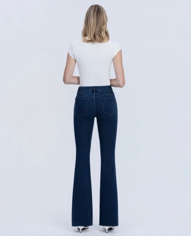A woman viewed from behind wearing a white short-sleeve top and dark blue flare jeans against a plain light gray studio background.