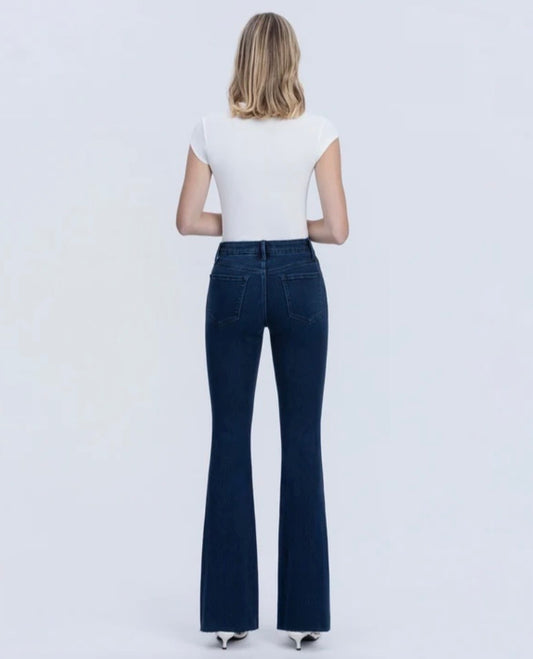 A woman viewed from behind wearing a white short-sleeve top and dark blue flare jeans against a plain light gray studio background.