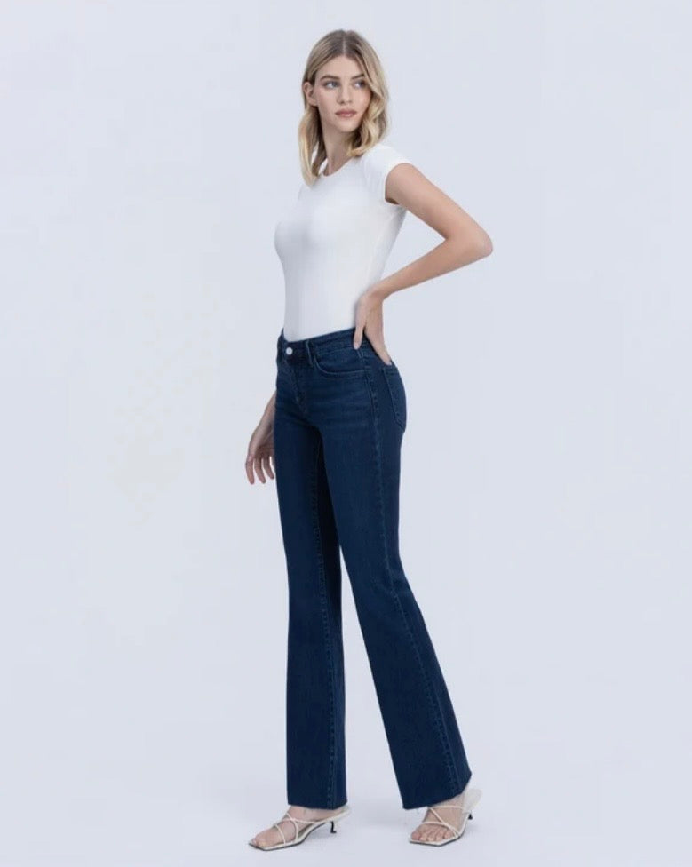 A woman modeling a pair of dark blue flare jeans with a white sleeveless top and beige heeled sandals, standing against a light gray studio background.